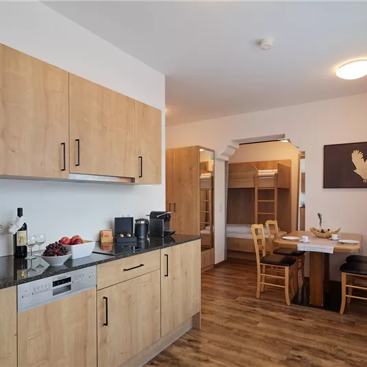 A modern kitchen with wooden cabinets and light walls. A dining table with chairs is visible in the background.