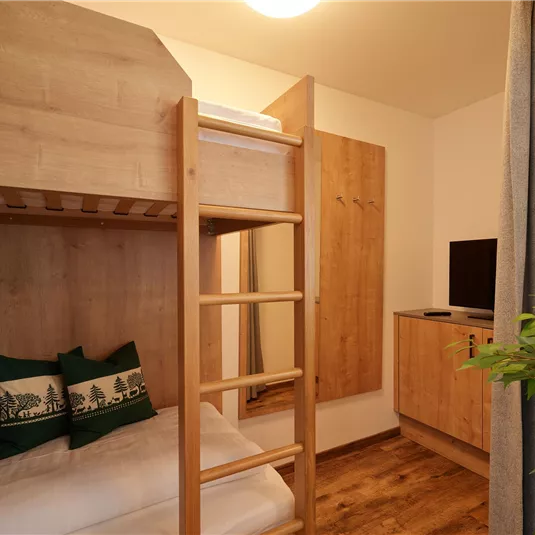 A modern bedroom with a wooden loft bed and cozy cushions. There is a television on the wall and a small wardrobe.