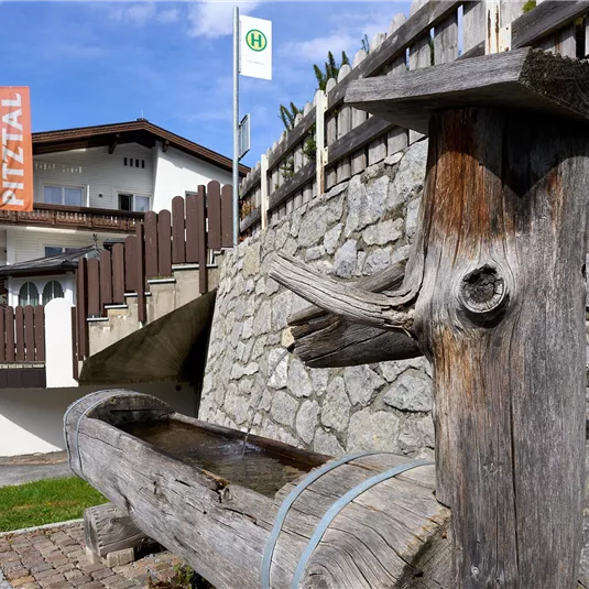 A wooden fountain stands in front of a stone house with a sign. In the background, green mountains and a clear sky are visible.