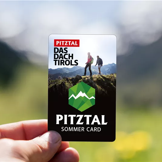 Eine Person hält eine Sommerkarte für das Pitztal in der Hand. Im Hintergrund sind Berge zu sehen, die eine schöne Naturkulisse bieten.