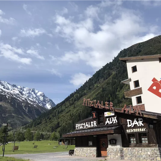 Ein charmantes Gebäude am Fuße der Berge mit einem Restaurant und einer Terrasse. Im Hintergrund sind schneebedeckte Gipfel und grüne Wiesen zu sehen.