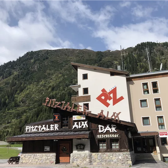 Ein modernes Gebäude mit einem traditionellen Gasthaus im Vordergrund. Im Hintergrund sind grüne Berge zu sehen.