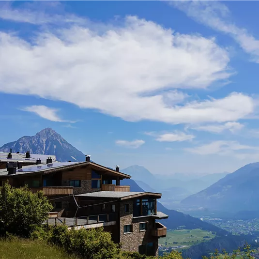 Ein malerisches Bergpanorama mit einem modernen Almhüttenhaus im Vordergrund. Der Himmel ist klar und blau, mit einigen Wolken und majestätischen Bergen im Hintergrund.