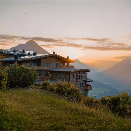 Eine malerische Berghütte mit Blick auf die umliegenden Berge. Im Hintergrund leuchtet der Sonnenuntergang über dem Tal.
