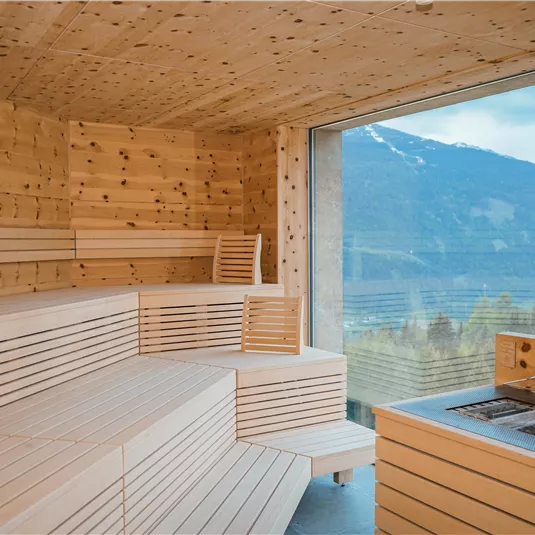 Ein modernes Saunainneres aus Holz mit großzügigen Bänken. Durch große Fenster bietet sich ein schöner Blick auf die Berge.