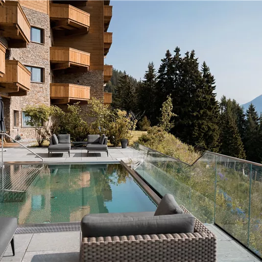 Eine moderne Terrasse mit einem Pool und gemütlichen Sitzgelegenheiten. Im Hintergrund sind Berge und Wälder sichtbar, die für eine entspannte Atmosphäre sorgen.