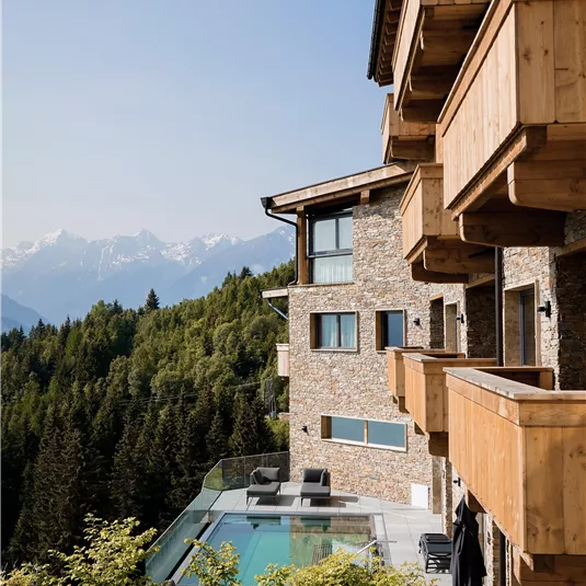 Ein modernes Gebäude im Gebirge mit Balkonen aus Holz. Im Vordergrund befindet sich ein Pool und eine grüne Landschaft.