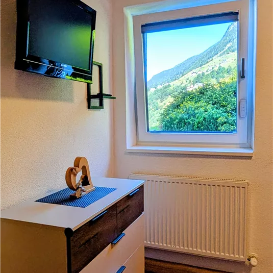A cozy room with a television on the wall and a window view of the Pitztal mountains. Under the window is a dresser with a decorative figure.