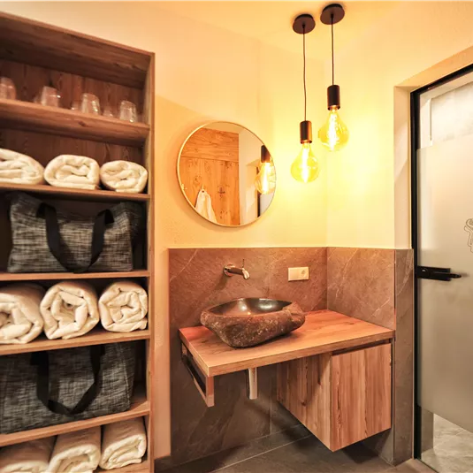 A stylish bathroom with wooden furniture and a modern washing facility. Folded towels and glasses are neatly arranged on a shelf.