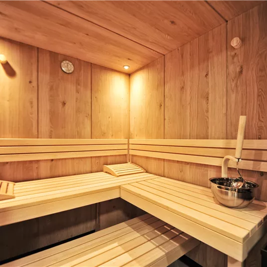A cozy sauna with wooden paneling and warm light. On one of the benches, there is a bowl of water and a wooden spoon.
