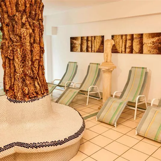 Ein entspannender Wellnessbereich mit Liegen und einem großen Baum in der Mitte. Die Wände sind mit ruhigen Naturmotiven dekoriert.