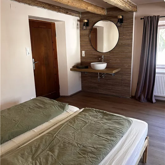 A cozy bedroom with two beds and green bed sheets. The wooden beams and the round mirror give the room a modern touch.