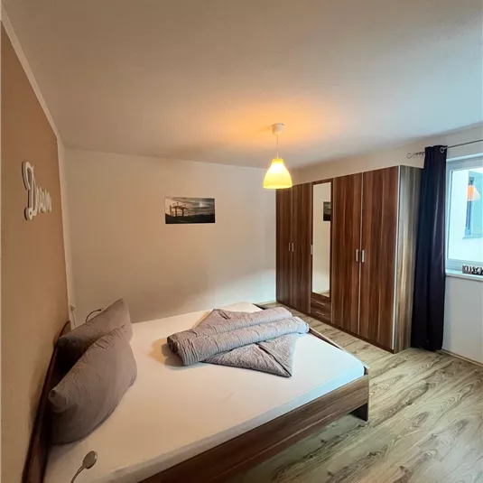 A modern bedroom with a double bed and wooden floor. The room has a wardrobe and plenty of natural light.