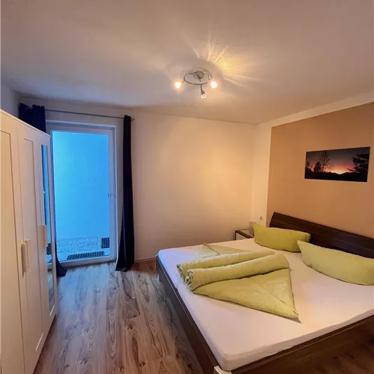 A simple bedroom with a double bed, light pillows, and a large window. The floor is made of wood and there is a closet.