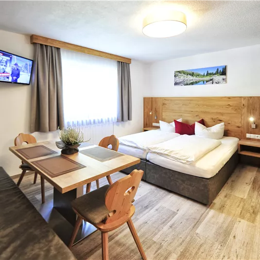 Ein modernes Zimmer mit einem Doppelbett, einem Tisch und Stühlen. Ein Fernseher und ein Fenster sorgen für Licht und Komfort.