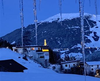 Eine verschneite Landschaft mit einem beleuchteten Haus unterhalb eines Berges. Der Himmel ist blau und es gibt Eiszapfen im Vordergrund.