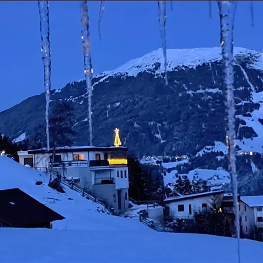 A snowy landscape with a illuminated house beneath a mountain. The sky is blue and there are icicles in the foreground.