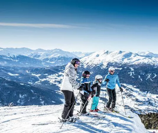 A group of four skiers stands on a snow-covered summit. In the background, majestic mountains and a clear blue sky can be seen.