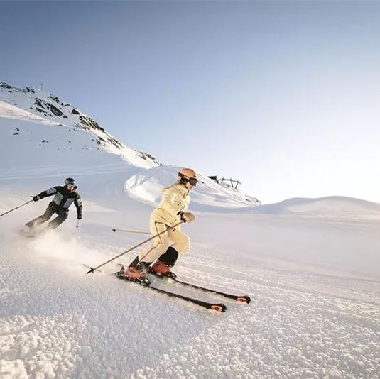 Two skiers are gliding through fresh, white snow in the mountains. The sky is clear and the sun is shining.