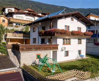 Ein malerisches Haus in einer bergigen Umgebung mit einem schönen Garten. Die Fassade ist hell und freundlich, umgeben von weiteren Wohnhäusern.