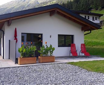 Ein modernes Haus inmitten grüner Hügel. Vor der Terrasse stehen blühende Pflanzen und zwei rote Stühle.