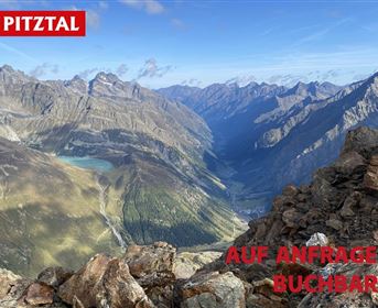 An impressive mountain landscape in the Pitztal with high peaks and a clear sky. In the foreground, rocks are visible, while in the valley green meadows and a lake can be seen.