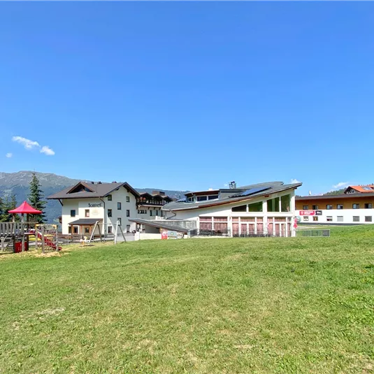 Ein weitläufiges Gelände mit mehreren Gebäuden und einem Spielplatz. Der Himmel ist klar und blau, umgeben von Bergen und grünen Wiesen.
