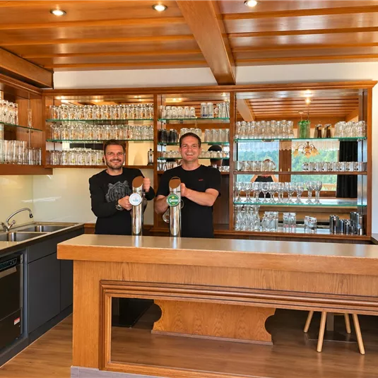 Zwei lächelnde Bartender stehen hinter einer Theke in einem einladenden Barbereich. Die Regale sind mit Gläsern und Getränken gefüllt.