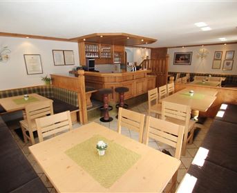 Ein gemütlicher Innenraum eines Lokals mit Holztischen und Bänken. Im Hintergrund befindet sich eine Theke mit Getränken und Beleuchtung.