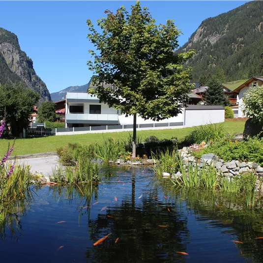 Ein ruhiger Teich mit goldenen Fischen und grünen Pflanzen. Im Hintergrund sieht man Berge und ein gemütliches Gebäude.
