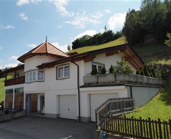 Ein modernes Haus mit einem roten Dach und einer Veranda. Es befindet sich in einer hügeligen Landschaft mit Bäumen im Hintergrund.