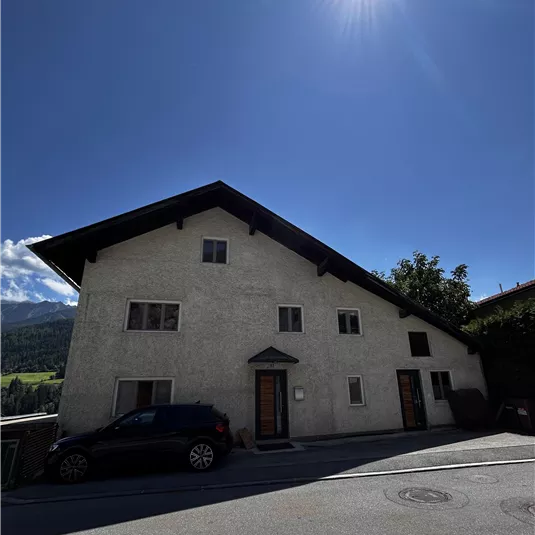 A two-story house with a bright facade and a black car in front of it. The sky is clear and sunny, surrounded by green mountains.