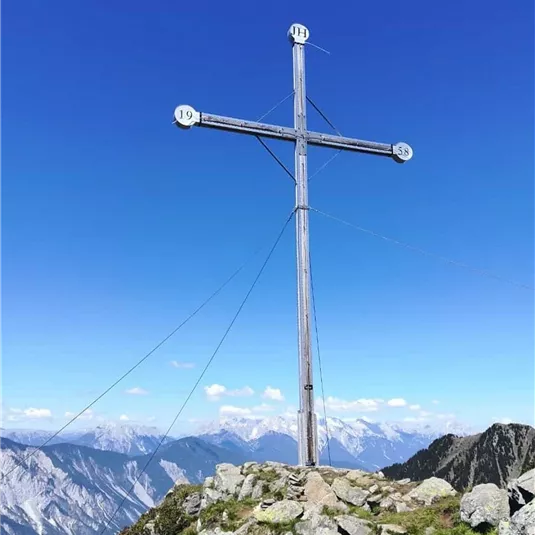 Ein Holzkreuz auf einem Berggipfel, umgeben von Felsen. Der Himmel ist klar und blau, mit Blick auf die umliegenden Berge.