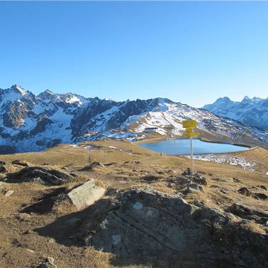 An impressive mountain landscape with snow-covered peaks and a calm lake in the foreground. The sky is clear and blue.