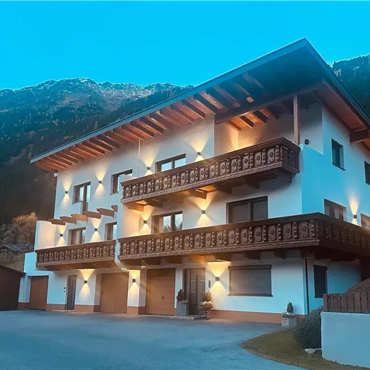 A modern house with several balconies and beautiful wood decor. In the background, mountains and a blue sky can be seen.