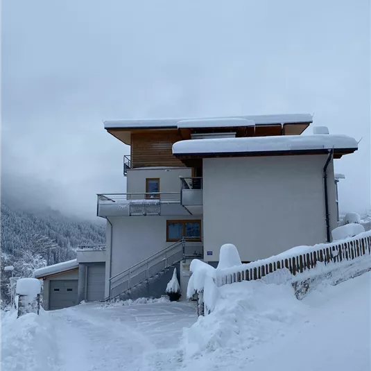 A modern house in the snow with a snow-covered driveway. The sky is overcast and the surroundings are wintry.