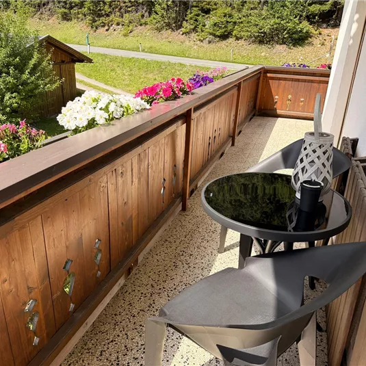 A cozy balcony with two chairs and a round table. Colorful flowers and a green landscape can be seen in the background.