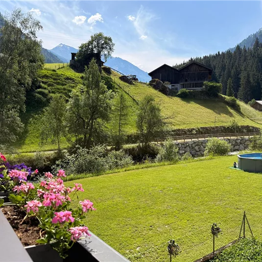 A green meadow with colorful flowers and a small pool. In the background, there are mountains and traditional huts.
