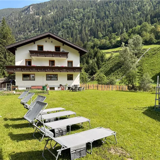 A cozy house in the mountains, surrounded by a lot of greenery. In the foreground, there are loungers on a well-kept lawn.