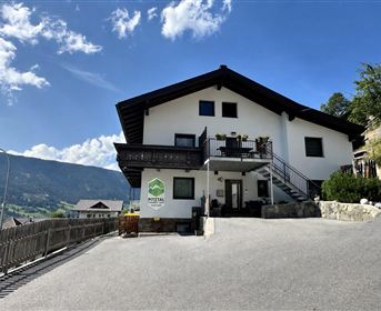 A modern house with a spacious terrace and a clear sky in the background. The surrounding landscape is characterized by mountains and greenery.