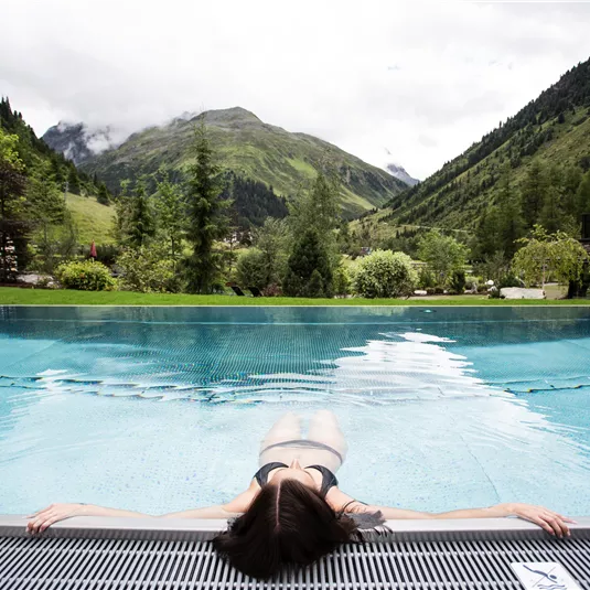 Ein entspannender Pool mit Blick auf die Berge. Eine Person liegt relaxed am Rand des Pools und genießt die Natur.