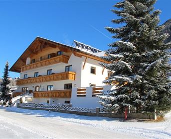 A charming house in the snow with a wooden-clad balcony.
Surrounded by snow-covered fir trees and a clear blue sky.
