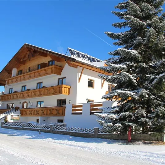 A charming house in the snow with a wooden-clad balcony.  
Surrounded by snow-covered fir trees and a clear blue sky.