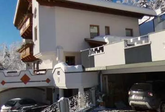A modern house in the snow, surrounded by snowy trees and mountains. The driveway is equipped with vehicles and the sun is shining.