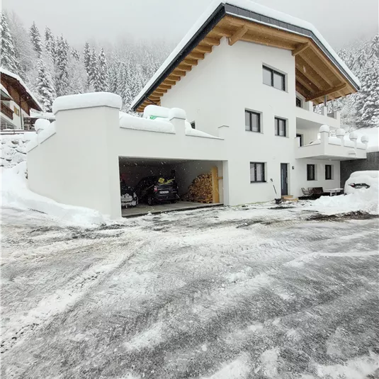 A modern house in the snow with a large garage area.  
Surrounded by snow-covered trees and a wintry landscape.