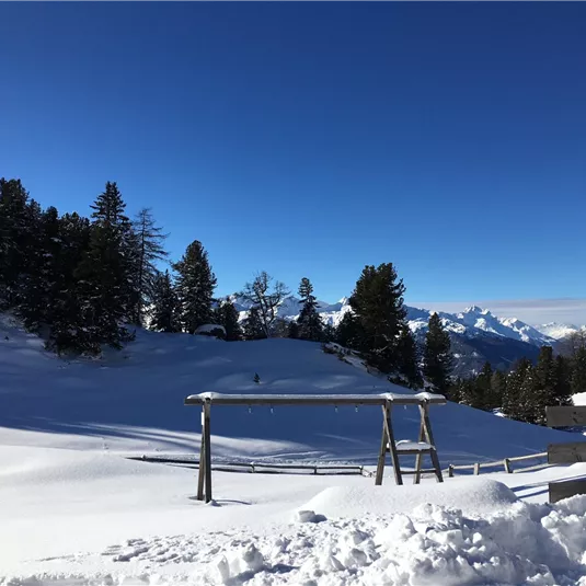 A snowy landscape with trees and a clear blue sky. In the foreground, there is a wooden swing.