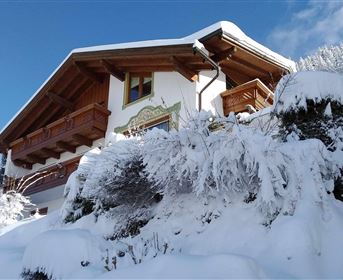 A charming chalet in the snow with a balcony and snow-covered trees. The sky is clear and blue.