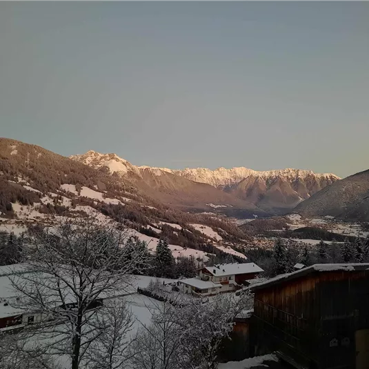 A winter mountain landscape with snow-covered hills and high peaks in the background. Small houses can be seen in the valleys.