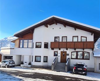 A modern, multi-story house with a clear blue sky in the background. The path in front of the house is covered with snow and several cars are parked in the parking lot.