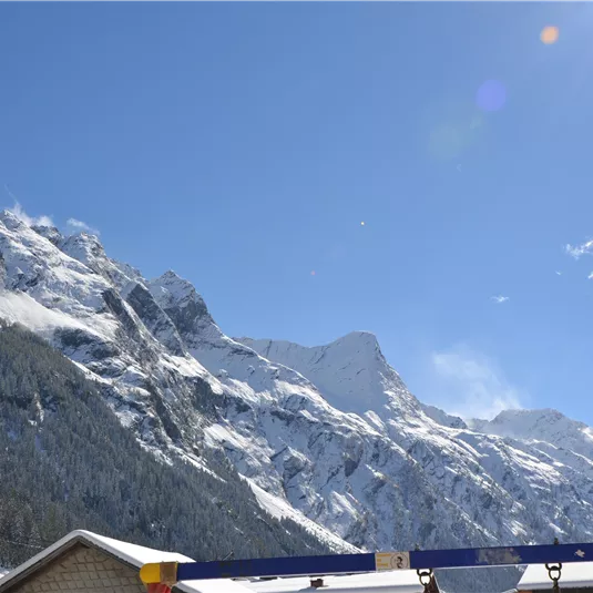 An impressive mountain landscape with snow-covered peaks under a clear blue sky. At the bottom, shadows of buildings can be seen.
